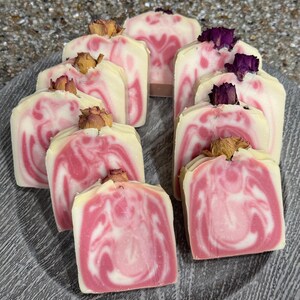 May include: A collection of handmade soaps with a swirl design in shades of pink and white, topped with dried rosebuds. The soaps are arranged on a gray, textured plate, showcasing their unique, artisanal appearance.