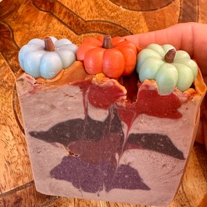 May include: A handmade soap bar with three decorative pumpkin accents in blue, orange, and green. The soap has a layered design with shades of red, brown, and purple. The soap is a fall-themed product.
