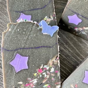 May include: Three bars of handmade soap with a dark gray base and purple star and butterfly designs. The soap is speckled with colorful flecks.
