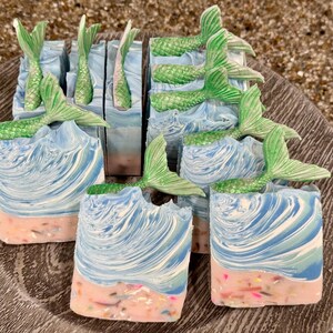 May include: Artisan soap bars with a mermaid theme. The soaps feature a beach scene with blue and white swirls representing waves, a pink sandy base, and green mermaid tails. The soaps are arranged on a decorative wooden plate.
