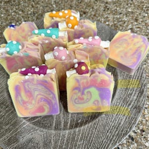 May include: A collection of colorful, swirled soaps in shades of yellow, pink, and purple, topped with decorative mushroom-shaped accents. The soaps are arranged on a gray, textured plate, creating a visually appealing display.