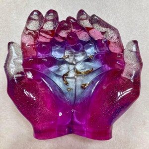 May include: A pair of hands sculpted in resin, with a gradient of pink, purple, and white, and flecks of glitter. The hands are cupped together, and there are flecks of gold leaf inside.