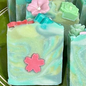 May include: A set of handmade soap bars with a floral theme. The bars are green, white, and yellow with pink and red flower accents.