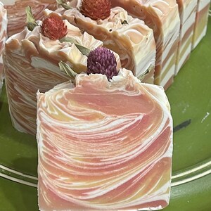 May include: Close-up of handmade soap bars with a swirled design in shades of peach, cream, and yellow. Each bar is topped with a dried flower and green leaves. The soap has a natural, organic appearance.