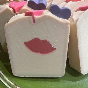 May include: Close-up of handmade soap bars with a romantic theme. The bars are off-white with a red lip design and topped with pink and purple heart-shaped embellishments. The soap is on a green surface.