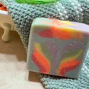 May include: A colorful, rectangular bar of soap with a marbled design. The soap features swirls of orange, yellow, red, and purple hues, set against a gray background. It rests on a textured, light green towel, suggesting a spa or bath product.