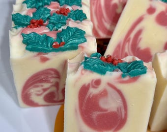 May include: Rectangular bars of handmade soap featuring a creamy white base with swirling pink patterns. The tops of the soap bars are decorated with molded green holly leaves and small red berries, creating a festive and detailed appearance.