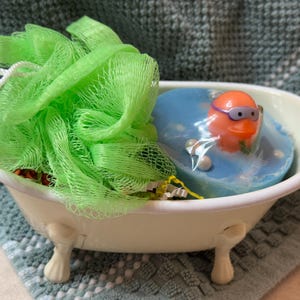 May include: A small, off-white bathtub-shaped container holds a green loofah and a blue soap with an orange rubber ducky wearing goggles. The tub sits on a textured, blue and gray surface.