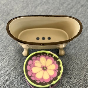 May include: A white ceramic bathtub soap dish with three drainage holes and a decorative floral soap bar with pink, white, and green swirls.