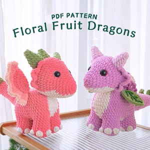 May include: Two crocheted plush dragon toys. One is pink with green leaves and the other is red with green leaves. The toys are sitting on a wooden table. PDF PATTERN Floral Fruit Dragons.