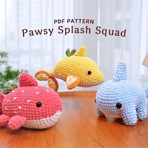 May include: Three crocheted plush toys shaped like fish. The fish are in different colors: pink, yellow, and blue. The text "Pawsy Splash Squad" is written above the toys.