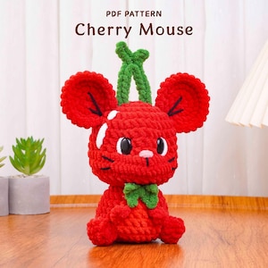 May include: A handmade red crochet mouse with large ears and a green stem, resembling a cherry. The mouse has white accents, a pink nose, and a green bow. The text "PDF PATTERN Cherry Mouse" is displayed at the top.