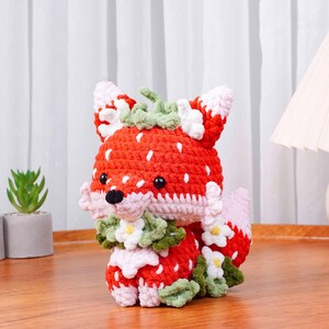 May include: A crocheted fox figurine with a strawberry design. The fox is primarily red with white speckles, resembling a strawberry. It has black eyes, a black nose, and is adorned with green leaves and white flowers. The fox is sitting on a wooden surface.