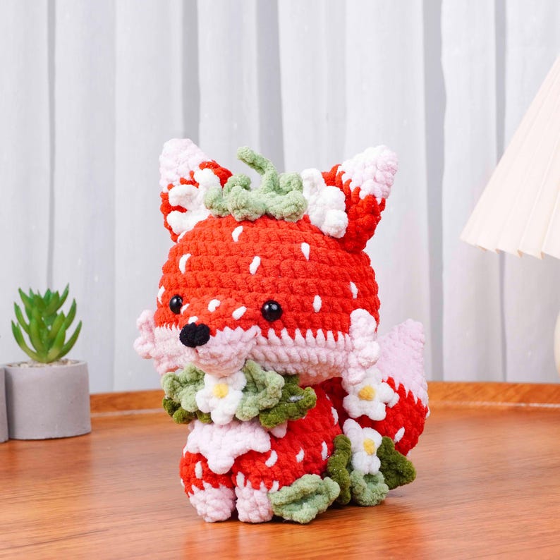 May include: A crocheted fox figurine with a strawberry design. The fox is primarily red with white speckles, resembling a strawberry. It has black eyes, a black nose, and is adorned with green leaves and white flowers. The fox is sitting on a wooden surface.