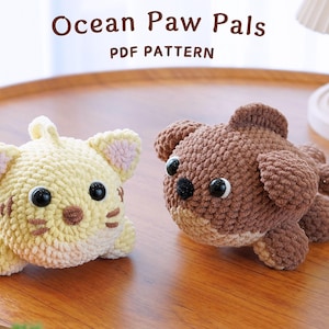 May include: Two crocheted stuffed animals, a yellow cat and a brown dog, on a wooden surface. The text "Ocean Paw Pals PDF PATTERN" is above the animals.