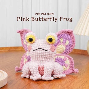 May include: A handmade crochet Pink Butterfly Frog. The frog is light pink with large butterfly wings in shades of pink and purple. The eyes are yellow with black pupils. The text "PDF PATTERN Pink Butterfly Frog" is above the frog.