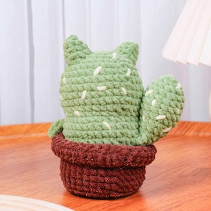 Op de afbeelding: Een handgemaakte, groene gehaakte cactus met witte accenten, in een bruine, getextureerde pot. De cactus heeft een unieke vorm met twee puntige oren en een zijarm. De zachte, pluche textuur voegt een gezellige, decoratieve touch toe.