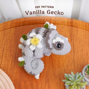 May include: A crocheted gecko in shades of gray and brown, adorned with white and yellow flower accents. The text "PDF Pattern Vanilla Gecko" is at the top. The gecko is on a wooden surface, with small potted plants in the background.
