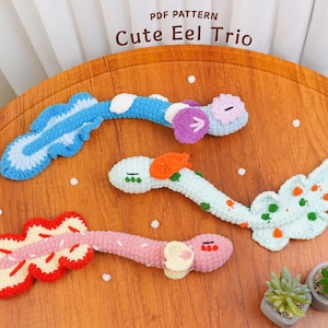 Pode incluir: Três brinquedos de enguia de crochê em azul, verde e rosa, com barbatanas e detalhes decorativos. O texto "Cute Eel Trio" é visível. Os brinquedos estão dispostos sobre uma superfície de madeira, com pequenas plantas em vasos em primeiro plano.