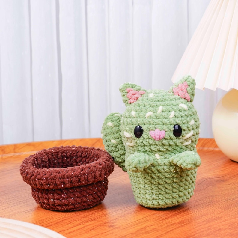 Op de afbeelding: Een handgemaakte gehaakte cactuskat en een bruine pot. De kat is groen met roze accenten en zwarte ogen. De pot is donkerbruin. De items staan op een houten oppervlak.