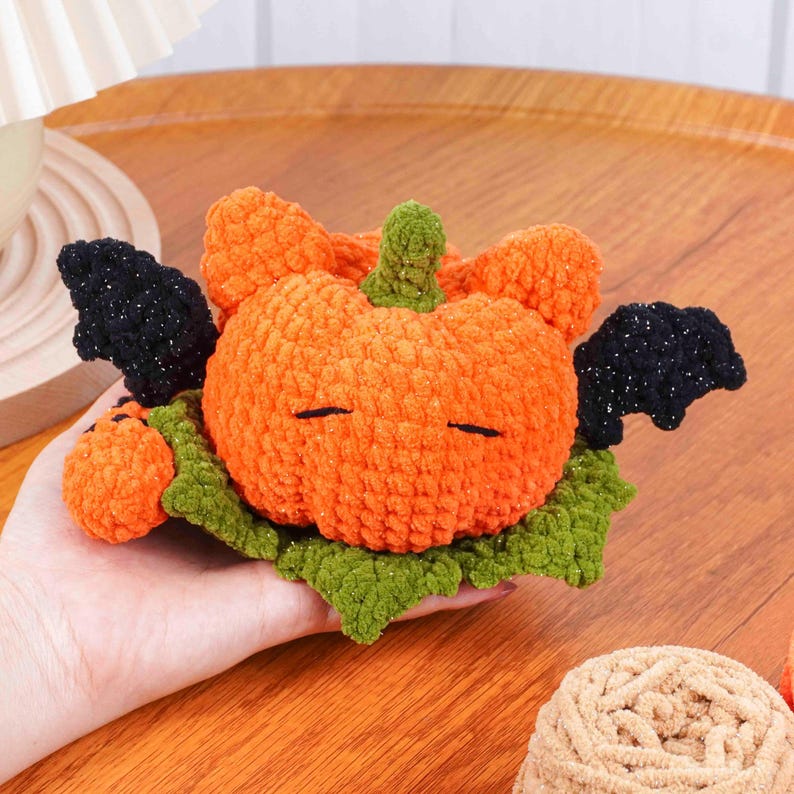May include: A handmade orange crochet pumpkin with black bat wings and a green stem. The pumpkin rests on a green leaf base, perfect for Halloween decor. The pumpkin has closed eyes.