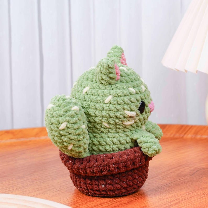 Op de afbeelding: Een handgemaakte gehaakte cactus knuffel in een terracotta pot. De cactus is lichtgroen met witte en roze accenten en zwarte ogen. De knuffel is gemaakt van zacht garen en is een schattig decoratief item.
