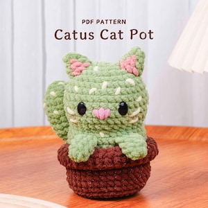 May include: A crocheted cat-shaped cactus pot in shades of green, brown, and pink. The cat has black eyes and a pink nose. The pot is brown and the text "PDF PATTERN Catus Cat Pot" is at the top.