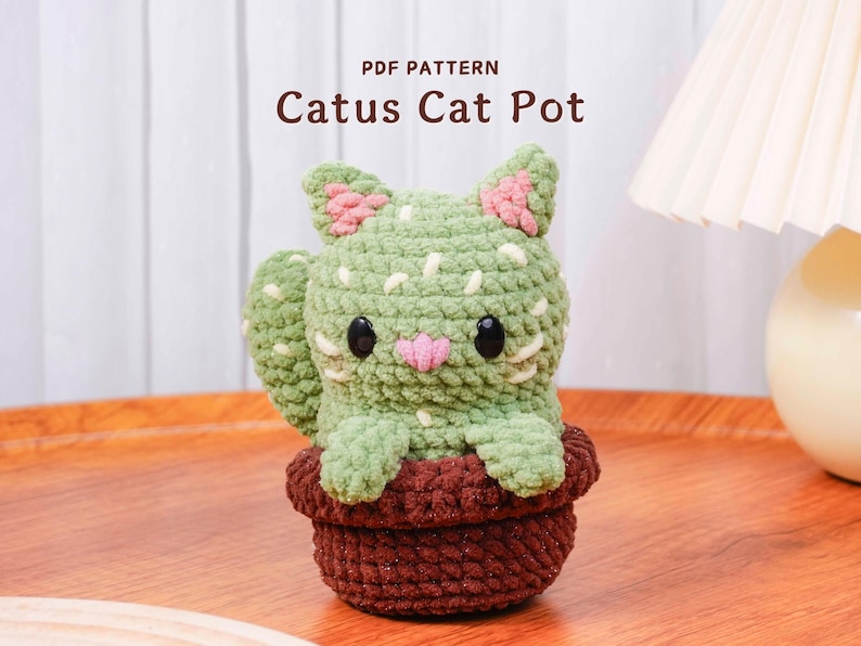 Op de afbeelding: Een gehaakte katvormige cactuspot in groen-, bruin- en roze tinten. De kat heeft zwarte ogen en een roze neus. De pot is bruin en de tekst "PDF PATTERN Catus Cat Pot" staat bovenaan.