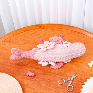 May include: A handmade, plush fish toy in shades of pink, decorated with white and pink flower accents. The fish has a tail and fins, and is made of a soft, textured material. A charming decorative item for a nursery or child's room.