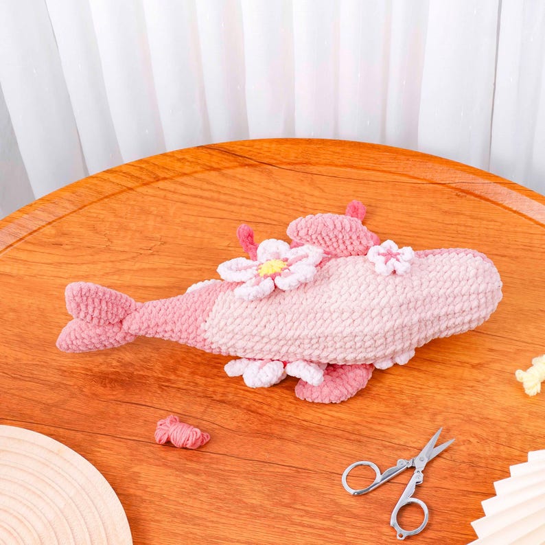 May include: A handmade, plush fish toy in shades of pink, decorated with white and pink flower accents. The fish has a tail and fins, and is made of a soft, textured material. A charming decorative item for a nursery or child's room.