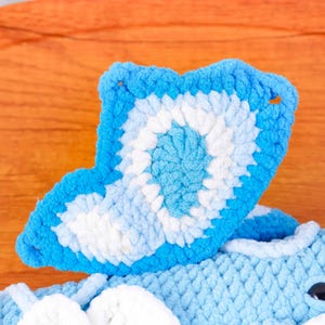 May include: A close-up shot of a blue and white crocheted item, possibly a toy or decorative piece. The item has a wing-like shape with a detailed pattern. The colour scheme includes shades of blue and white, with a textured appearance.