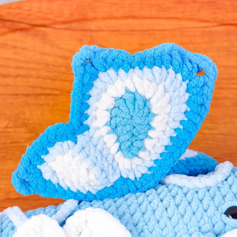 May include: A close-up shot of a blue and white crocheted item, possibly a toy or decorative piece. The item has a wing-like shape with a detailed pattern. The colour scheme includes shades of blue and white, with a textured appearance.