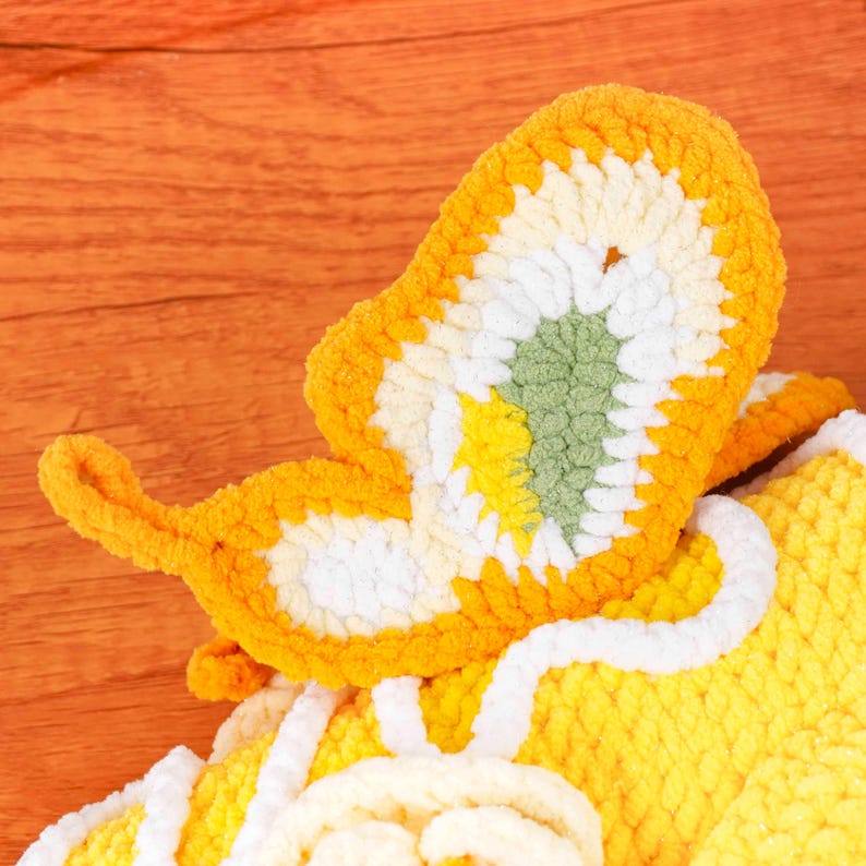 May include: A close-up of a handmade, crocheted butterfly in shades of yellow, white, and green. The butterfly has a textured, fluffy appearance, with detailed wing patterns. The background is a warm, wooden surface.