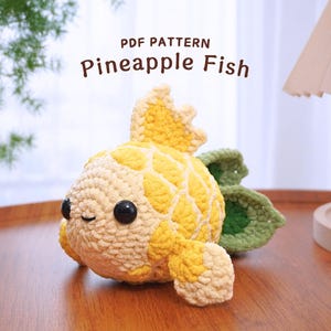 Peut inclure: Un jouet en crochet en forme de poisson ananas avec un corps jaune et blanc et des feuilles vertes. Le jouet a un visage mignon avec des yeux noirs et une bouche souriante. Le jouet est assis sur une surface en bois.