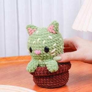Op de afbeelding: Een handgemaakte gehaakte kat in een pot. De kat is groen met roze accenten op de oren en neus, en zwarte ogen. De pot is bruin. De kat is ontworpen om op een cactus te lijken.