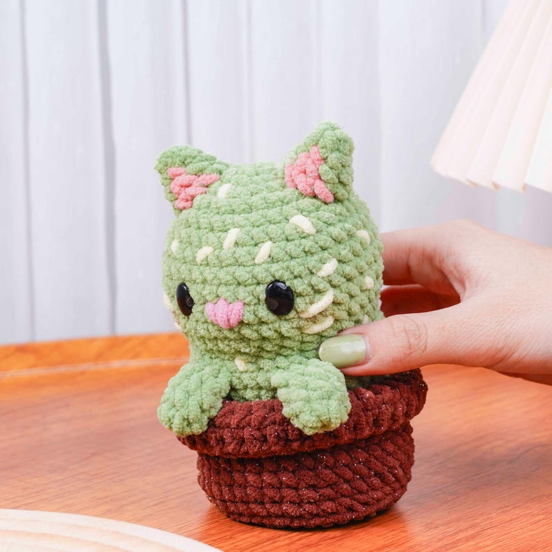 Op de afbeelding: Een handgemaakte gehaakte kat in een pot. De kat is groen met roze accenten op de oren en neus, en zwarte ogen. De pot is bruin. De kat is ontworpen om op een cactus te lijken.