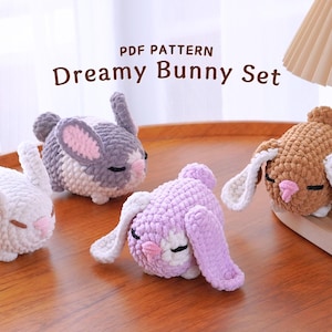 May include: A set of four crocheted bunny plushies in different colors, including white, gray, brown, and purple. The plushies have pink noses and closed eyes. The plushies are arranged on a wooden surface. The text "PDF PATTERN Dreamy Bunny Set" is displayed above the plushies.