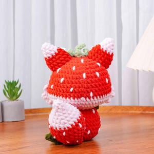 May include: A handmade crochet strawberry plush toy. The toy is primarily red with white seed details and pink and white accents. It has a green stem and leaves, and is sitting on a wooden surface.