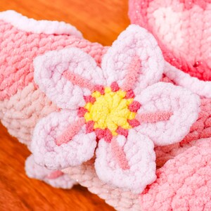 May include: A close-up of a handmade, plush, pink flower with a yellow centre and pink accents. The flower is attached to a soft, textured, pink and white fabric, possibly a scarf or blanket. The background is a warm, wooden surface.