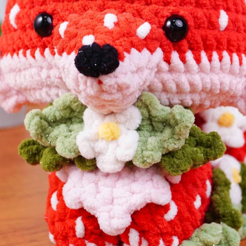 May include: A handmade crochet strawberry-themed plush toy. The toy features a red body with white spots, a pink face, and a green leafy collar with white flowers. The eyes are black and the nose is black.