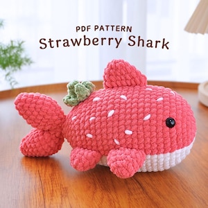 Pode incluir: Um brinquedo de pelúcia de tubarão morango de crochê com uma barriga branca e folhas verdes no topo da cabeça. O brinquedo é rosa e branco e tem um olho preto costurado.  PDF PATTERN Strawberry Shark