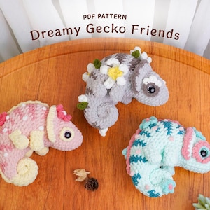 Könnte beinhalten: Drei gehäkelte Geckos in Pastellfarben: Rosa, Grau und Blau. Die Geckos haben florale Verzierungen und sind auf einer Holzoberfläche angeordnet. Der Text "Dreamy Gecko Friends" ist sichtbar.