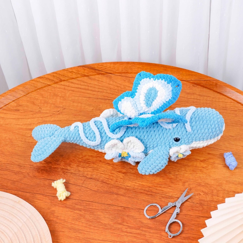 May include: A handmade blue crochet whale with a large butterfly wing design on its back. The whale has white accents and small white flowers. A pair of scissors and yarn scraps are on the wooden surface.