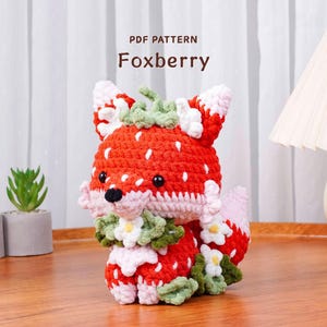 Puede incluir: Un juguete de peluche con forma de zorro tejido a ganchillo, con diseño de fresa. El zorro tiene pelaje rojo y blanco, ojos negros y una corona de hojas verdes. El texto "PDF PATTERN Foxberry" está encima del juguete.