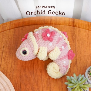Puede incluir: Un gecko tejido a ganchillo en tonos rosa y crema, adornado con detalles florales. El gecko está sobre una superficie de madera, con el texto "PDF PATTERN Orchid Gecko" visible arriba. Un artículo artesanal.