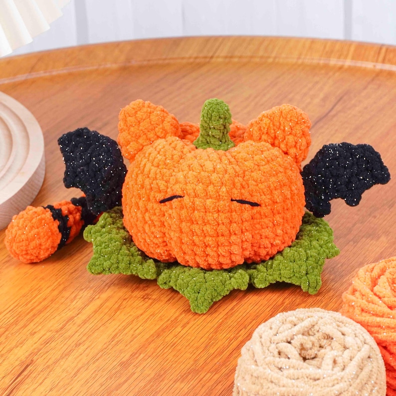 May include: A handmade orange crochet pumpkin with black glittery bat wings and a green stem, resting on green leaves. The pumpkin has two horizontal black lines for eyes. A Halloween-themed decorative item.