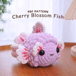 Pode incluir: Um brinquedo de pelúcia de crochê em forma de peixe com detalhes de flores de cerejeira rosa e branco. O peixe é roxo e tem olhos pretos. O brinquedo está sobre uma superfície de madeira. PDF PATTERN Cherry Blossom Fish