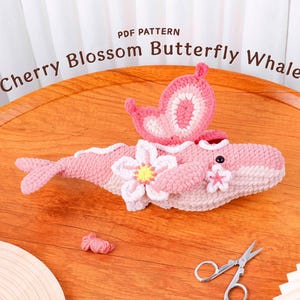 Pode incluir: Uma baleia rosa de crochê feita à mão com um design de borboleta e flor. A baleia tem um corpo rosa, detalhes brancos e uma flor em um lado. O texto "Cherry Blossom Butterfly Whale" está no topo.