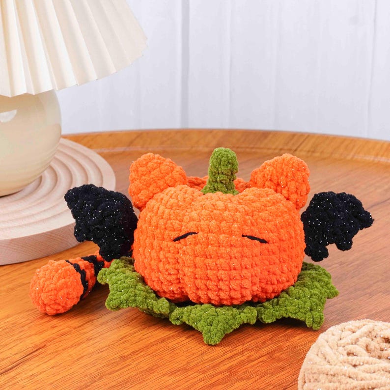 May include: A handmade orange crochet pumpkin with black glittery wings and a green stem and leaves. The pumpkin has closed eyes and is resting on a wooden surface. A Halloween-themed decorative item.