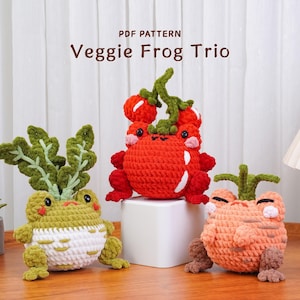 May include: Three crocheted frog planters in the shape of vegetables. One is green and white with leafy greens, another is red with tomato accents, and the third is orange with a carrot top. The text "PDF PATTERN Veggie Frog Trio" is at the top.